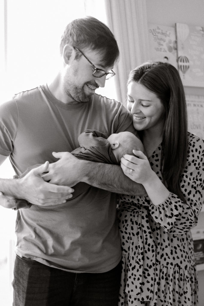 Black and white image of new parents holding their newborn son in front of large window.