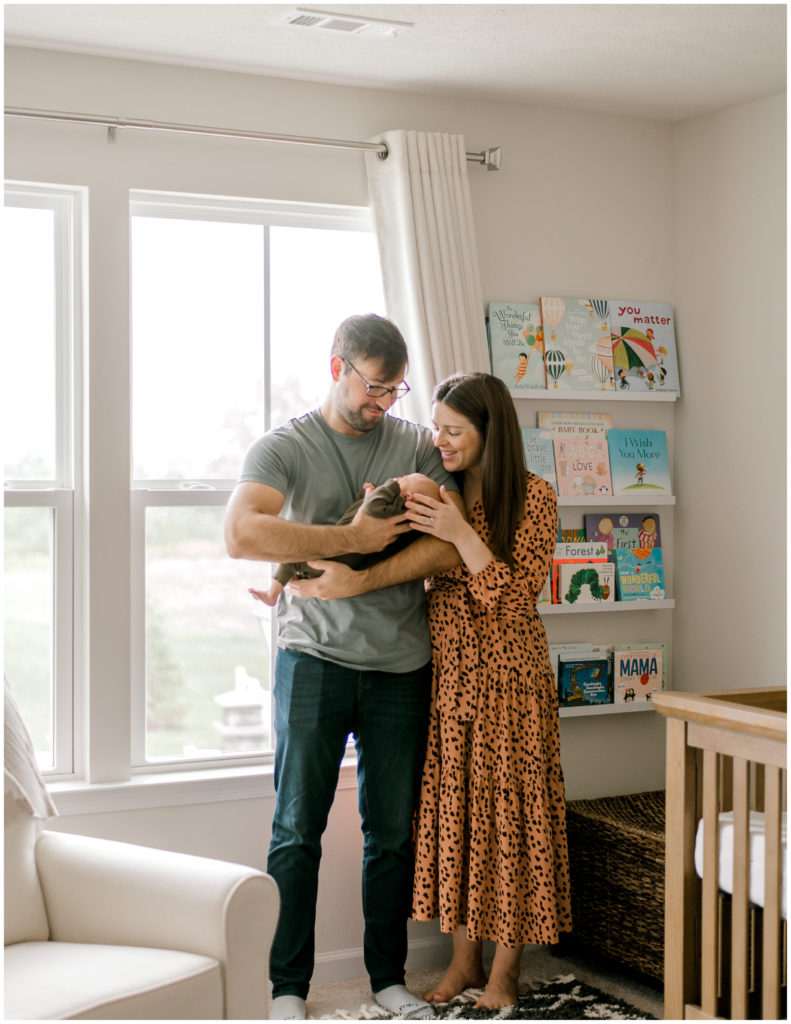 New mom and dad holding their newborn son in front of large window in neutral nursery.
