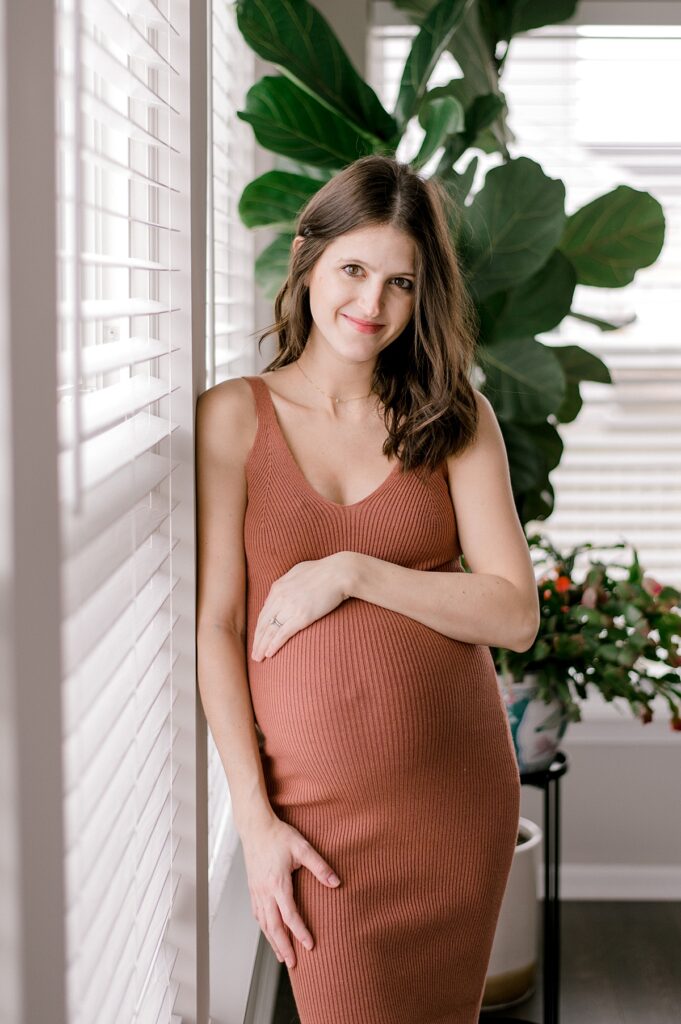 A smiling mom to be looking at the camera while holding her pregnant bump in a rust colored dress for a north ridgeville maternity session by maternity photographer, Brittany Serowski Photography.