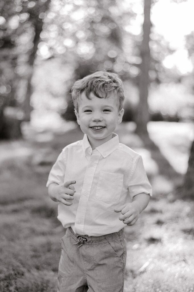 A toddler boy in a white button up shirt smiling at the camera in a black and white photo for a Schoepfle Gardens family session by Brittany Serowski Photography.