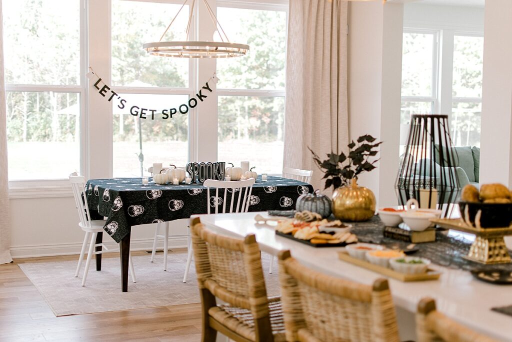 An eat in kitchen with a black and white skull tablecloth, topped with white pumpkand Halloween treats on the island for hosting a memorable Halloween. Party styling and design by One Stylish Party, Photographed by Brittany Serowski Photography.