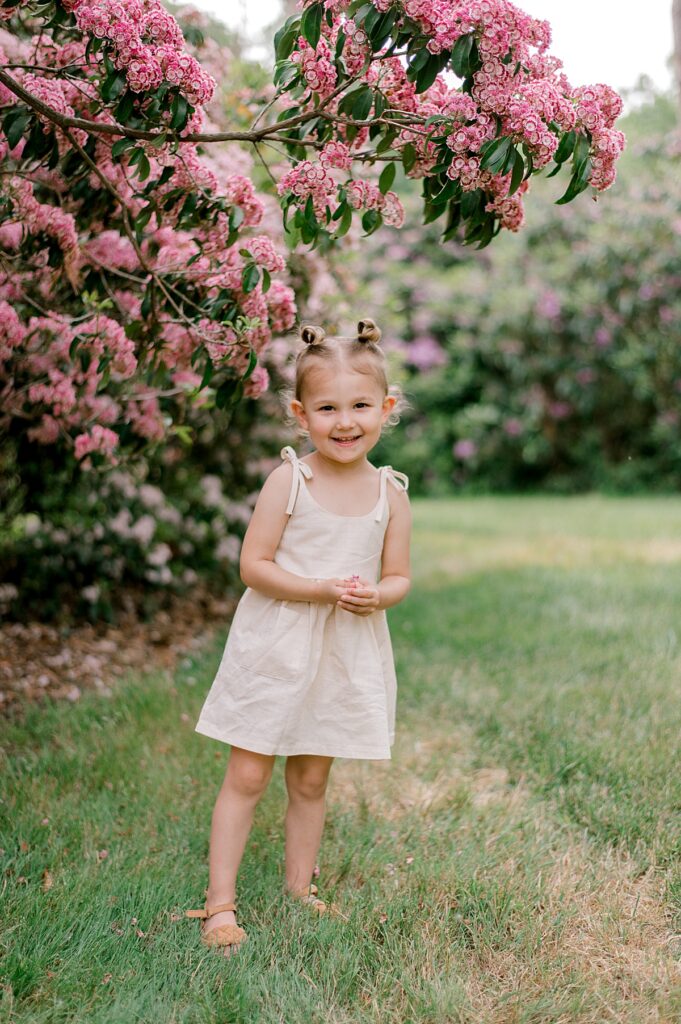 A little girl standing under a pink flowering bush at Schoepfle Gardens in a cream tie dress.