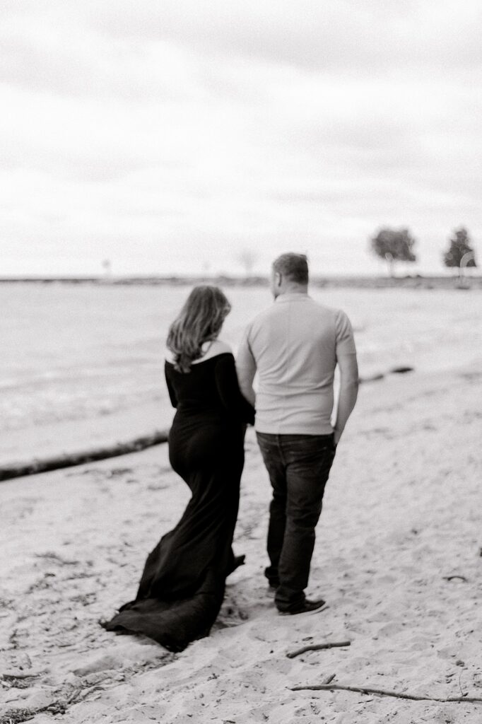 Newly expecting parents at Main Street Beach for a Vermilion beach maternity session on a cloudy & windy day. Mom is wearing a black, maxi dress and dad is wearing a gray polo and dark wash jeans. The couple is holding hands and walking away from the camera, along the shores of Lake Erie. Captured by photographer, Brittany Serowski Photography.