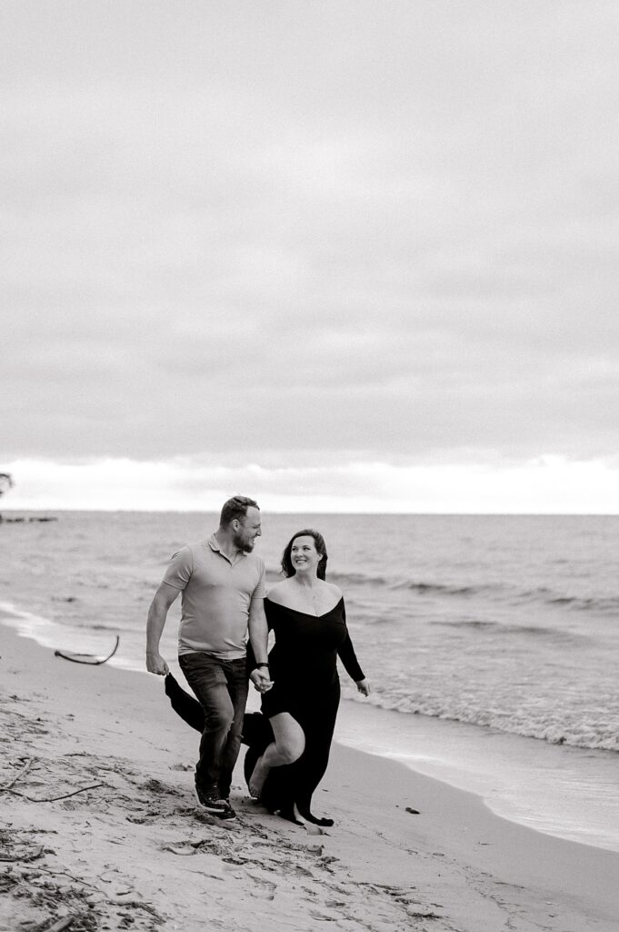 Newly expecting parents at Main Street Beach for a Vermilion beach maternity session on a cloudy & windy day. Mom is wearing a black, maxi dress and dad is wearing a gray polo and dark wash jeans. The expecting parents are running along the shoreline and smiling at one another while hand in hand. Captured by photographer, Brittany Serowski Photography.