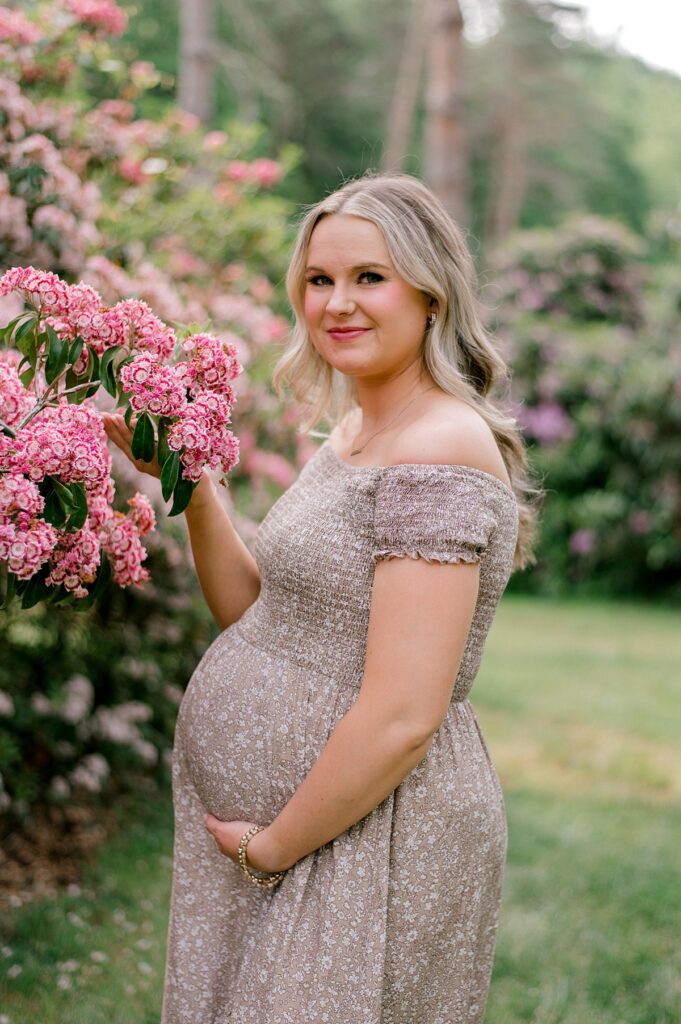 A Schoepfle Gardens maternity session by Brittany Serowski Photography of an expecting mother holding her belly & some pink flowers while standing next to a pink flowering tree.