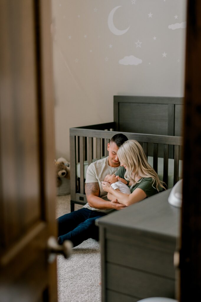 A new mom and dad sitting on the floor of a nursery in front of the crib, snuggle d by Cleveland Newborn Photographer, Brittany Serowski Photography.