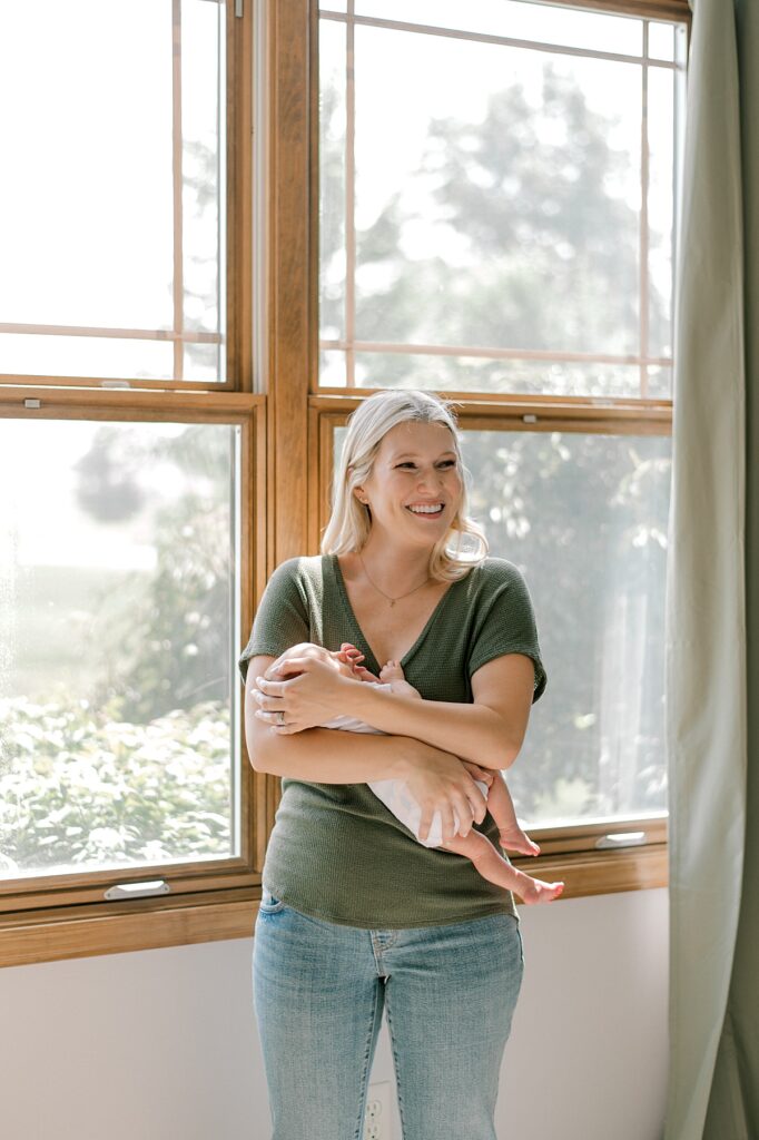 A new mom holding her newborn son in front of a large window in a green shirt & smiling off into the distance by Cleveland Newborn Photographer, Brittany Serowski Photography.