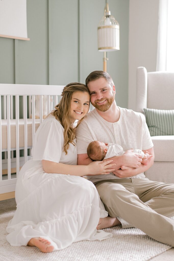 A Medina neutral nursery session by newborn photographer, Brittany Serowski Photography of a mother and father holding their newborn son smiling at the camera and sitting in front of his crib.