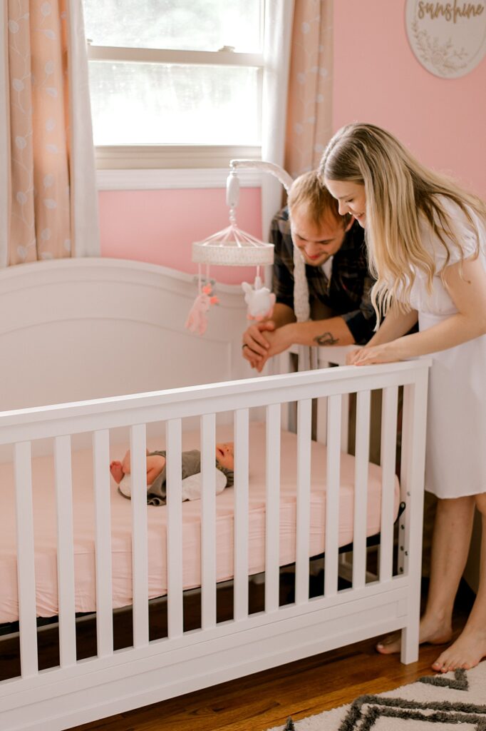 A mom and dad peering over the crib at the newborn daughter in her pink nursery and white crib by Parma newborn photographer, Brittany Serowski Photography.
