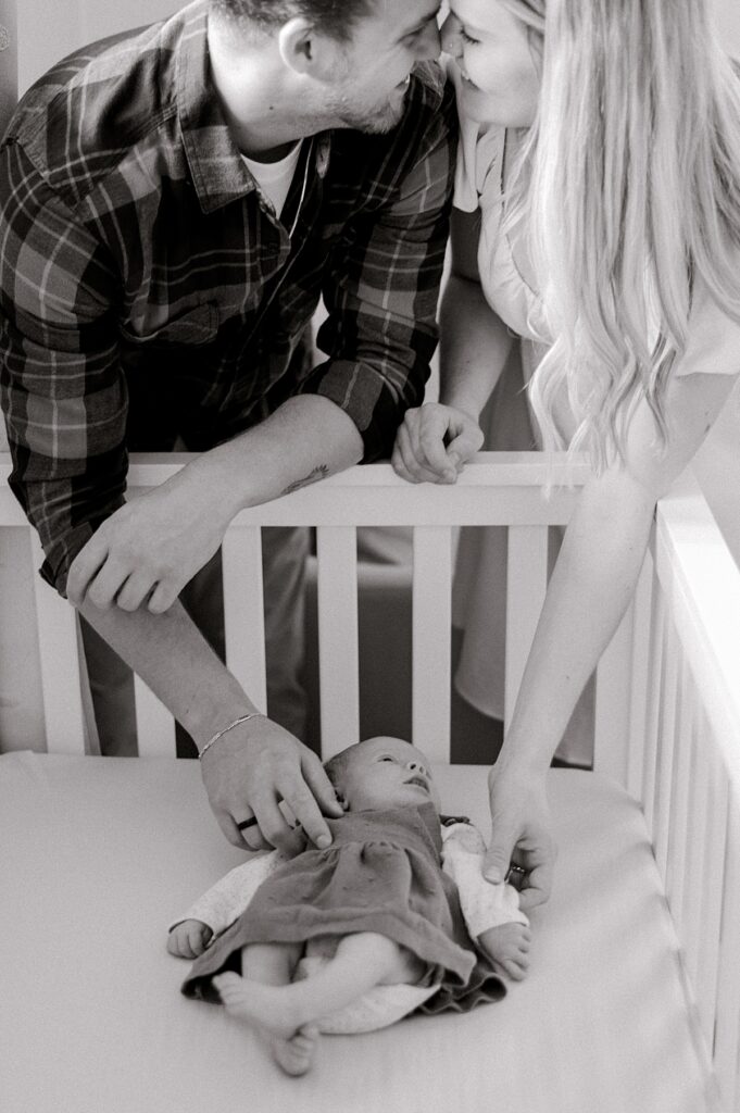 A mom and dad leaning over the crib of their newborn daughter while smiling at one another nose to nose, while their baby girl stares up at them by Parma newborn photographer, Brittany Serowski Photography.