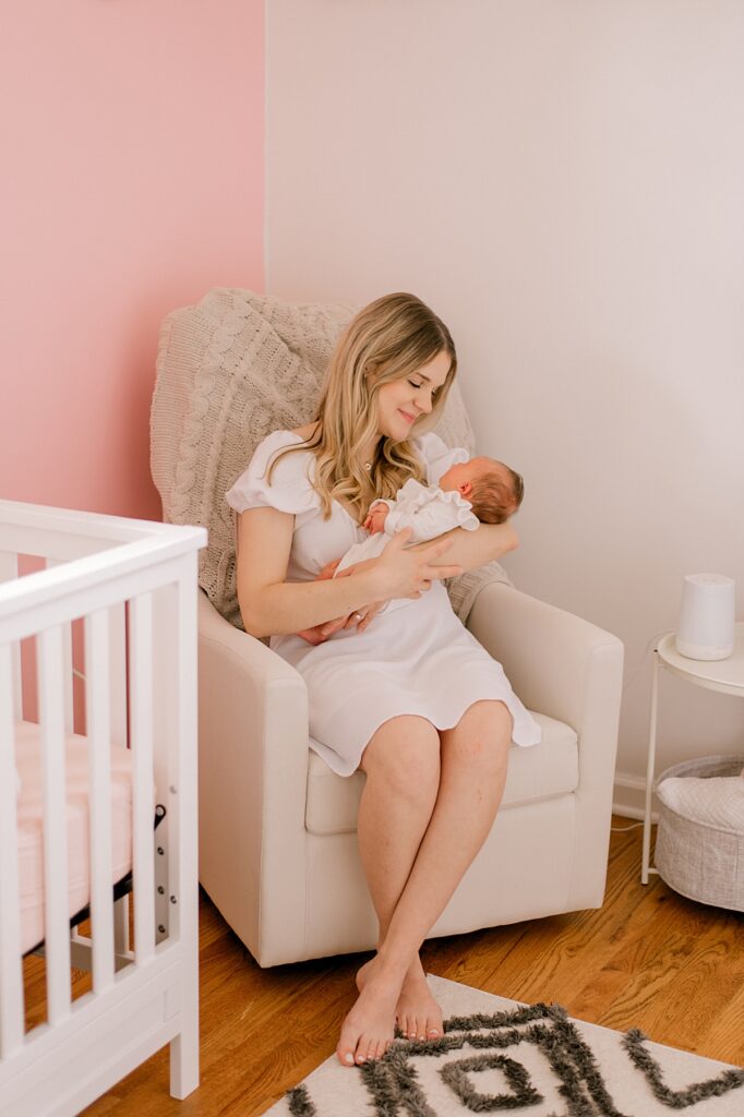 A mom sitting in a neutral beige rocking chair in her newborn daughter's pink nursery, while holding her daughter and staring at her by Parma newborn photographer, Brittany Serowski Photography.