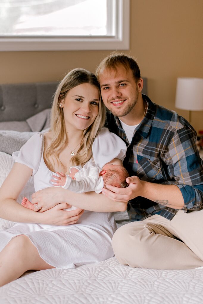 A mom and dad looking at the camera and smiling while sitting on their bed and holding their newborn daughter close by Parma newborn photographer, Brittany Serowski Photography.