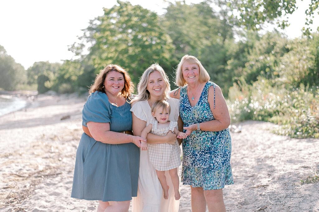 A three generations photo of a mom, her daughter and infant granddaughter in blue and neutral clothing and dresses along the shores of Lake Erie, smiling at the camera while snuggled close together by Cleveland Family Photographer, Brittany Serowski Photography.