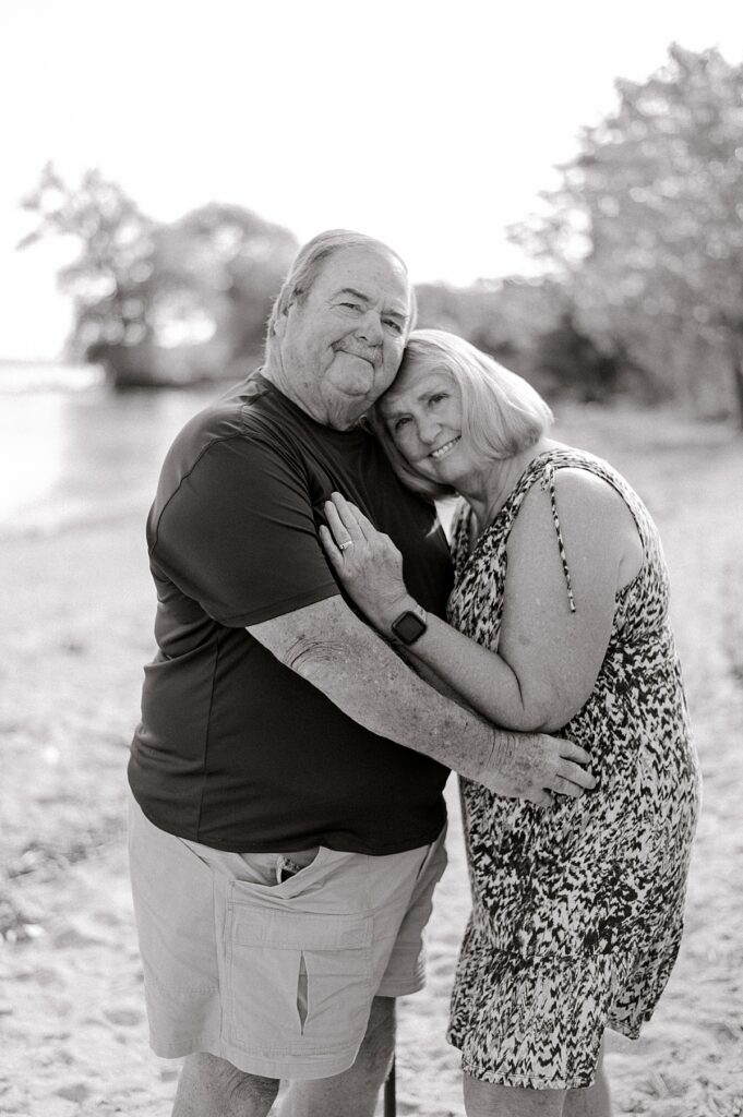 A couple along the shores of Lake Erie standing next to one another in blue and white clothing with a hand on his chest by Cleveland Family Photographer, Brittany Serowski Photography.