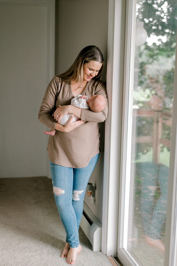 A mom leaning up against a sliding door window, holding her newborn son in a brown shirt and ripped jeans while gazing at her baby by Vermilion Newborn Photographer, Brittany Serowski Photography.