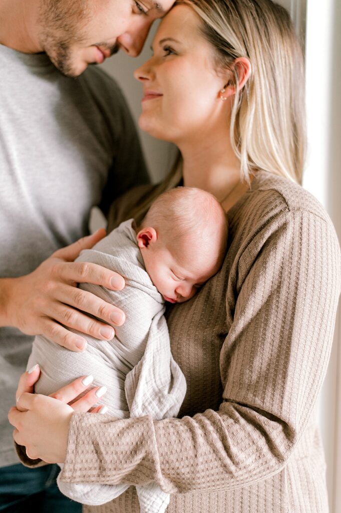 A mom and dad forehead to forehead, leaning on a large window while mom holds onto their newborn son by Vermilion Newborn Photographer, Brittany Serowski Photography.