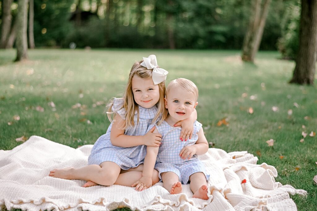 A young brother and sister duo in matching blue and white gingham summer outfits as they sit on a cream colored blanket, nestled together smiling at the camera by Chagrin Falls Family Photographer, Brittany Serowski Photography.