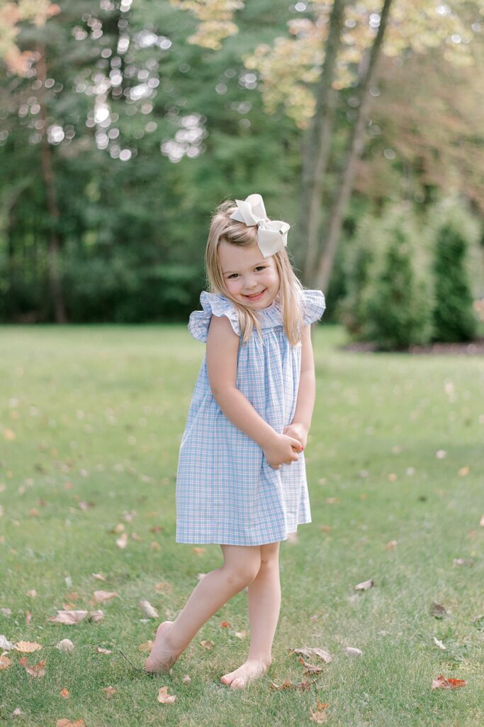 A little girl in blue and white gingham summer outfit and white bow in her hair, standing with her hands folded smiling at the camera by Chagrin Falls Family Photographer, Brittany Serowski Photography.