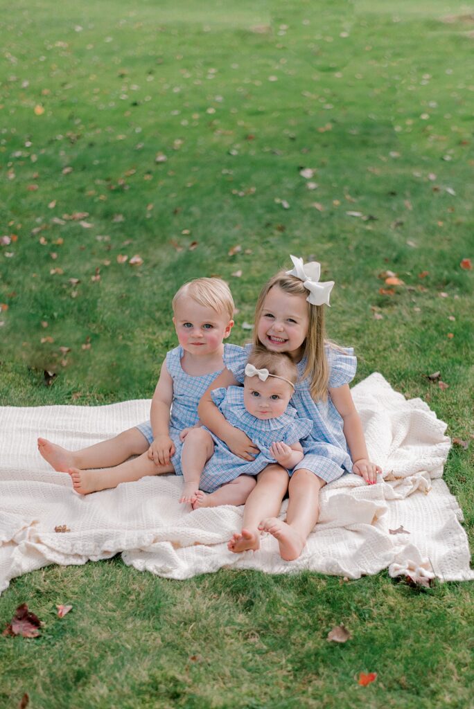 A group of three young siblings in matching blue and white gingham summer outfits as they sit on a cream colored blanket, nestled together smiling at the camera by Chagrin Falls Family Photographer, Brittany Serowski Photography.