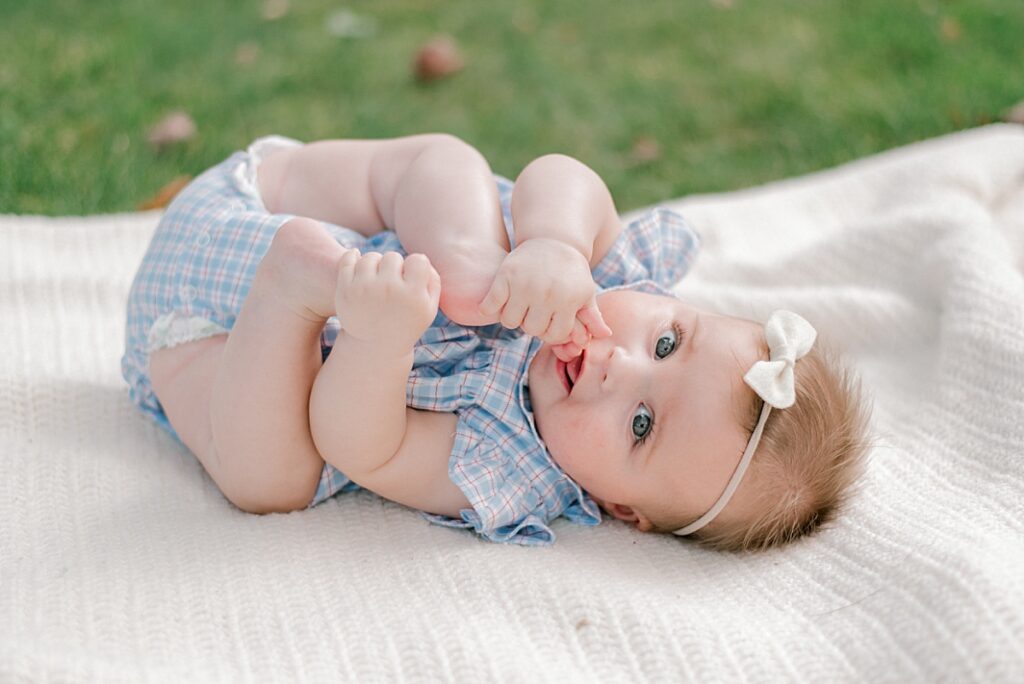 A young baby girl laying on a cream blanket eating her toes with a blue and white gingham dress on and white headband, looking at Chagrin Falls Family Photographer, Brittany Serowski Photography.