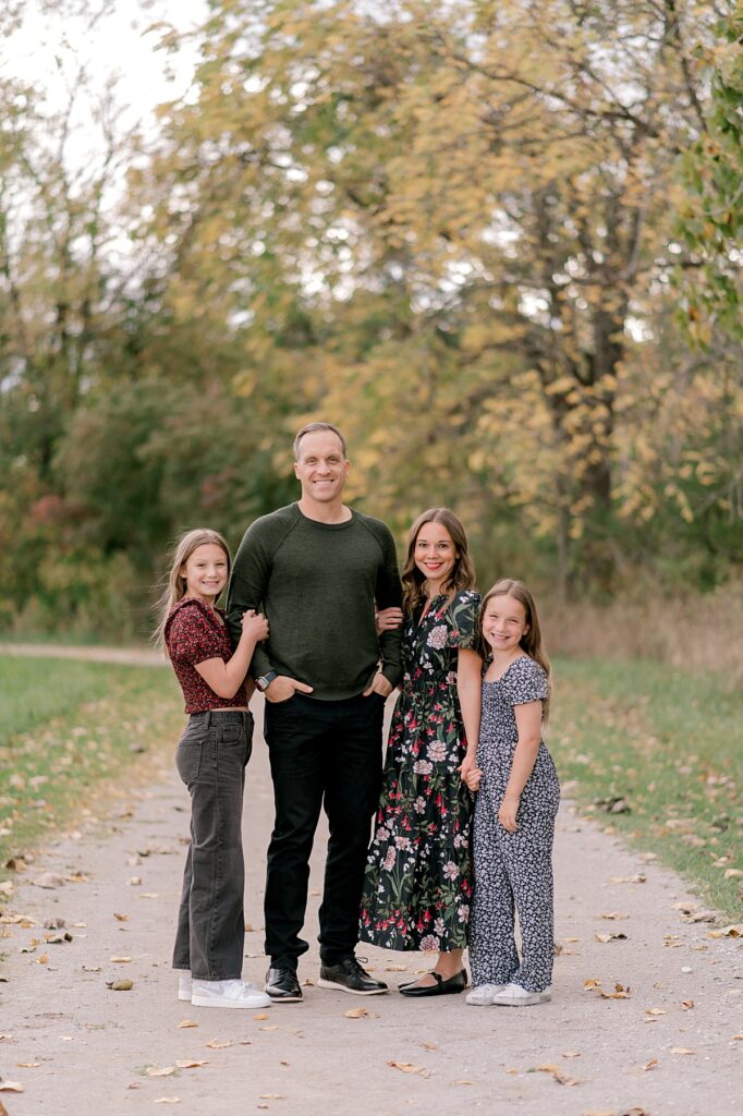 A family of four with two young daughters, in dark moody clothing smiling at the camera and standing close together while holding onto one another, by Cleveland fall family photographer, Brittany Serowski Photography.