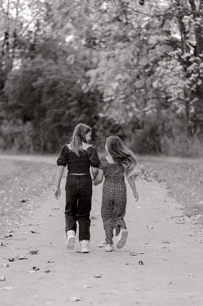 Two young sisters holding hands and running away from the camera down a gravel path in a black and white picture by Cleveland fall family photographer, Brittany Serowski Photography.