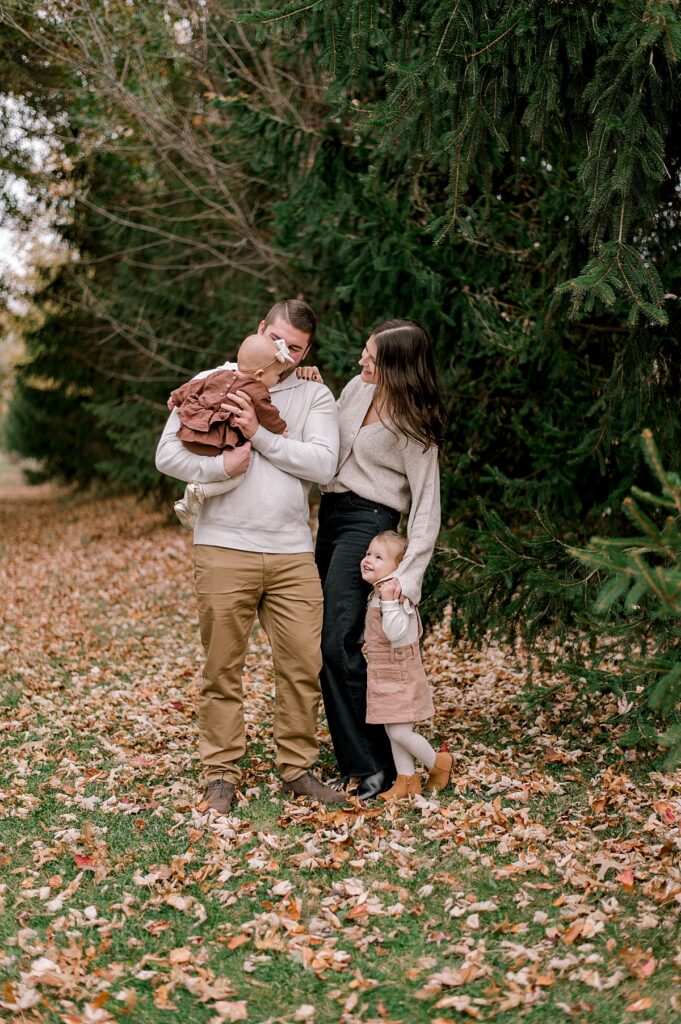 A family of four in all neutral colored clothes standing in front a row of pine trees while smiling at one another and laughing by Cleveland fall family photographer, Brittany Serowski Photography.