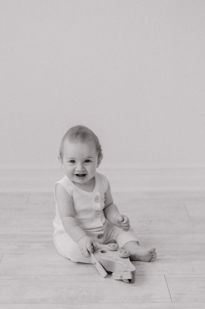A black and white portrait of a 6 month old baby in a neutral romper with wood buttons in front of an all white background at Lake Arts Studio, smiling at the camera and playing with a wooden airplane by Cleveland Personality Portraits photographer, Brittany Serowski Photography.