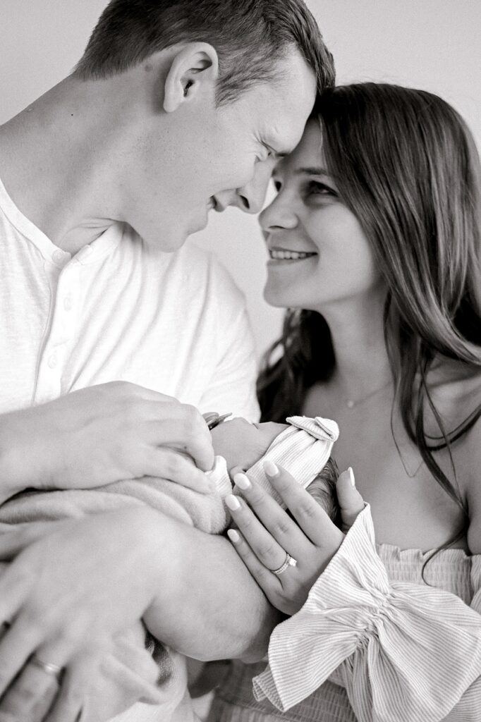 A black and white image of a mom and dad, forehead to forehead smiling at one another while holding close their newborn baby daughter, by Cleveland Newborn Photographer, Brittany Serowski Photography.