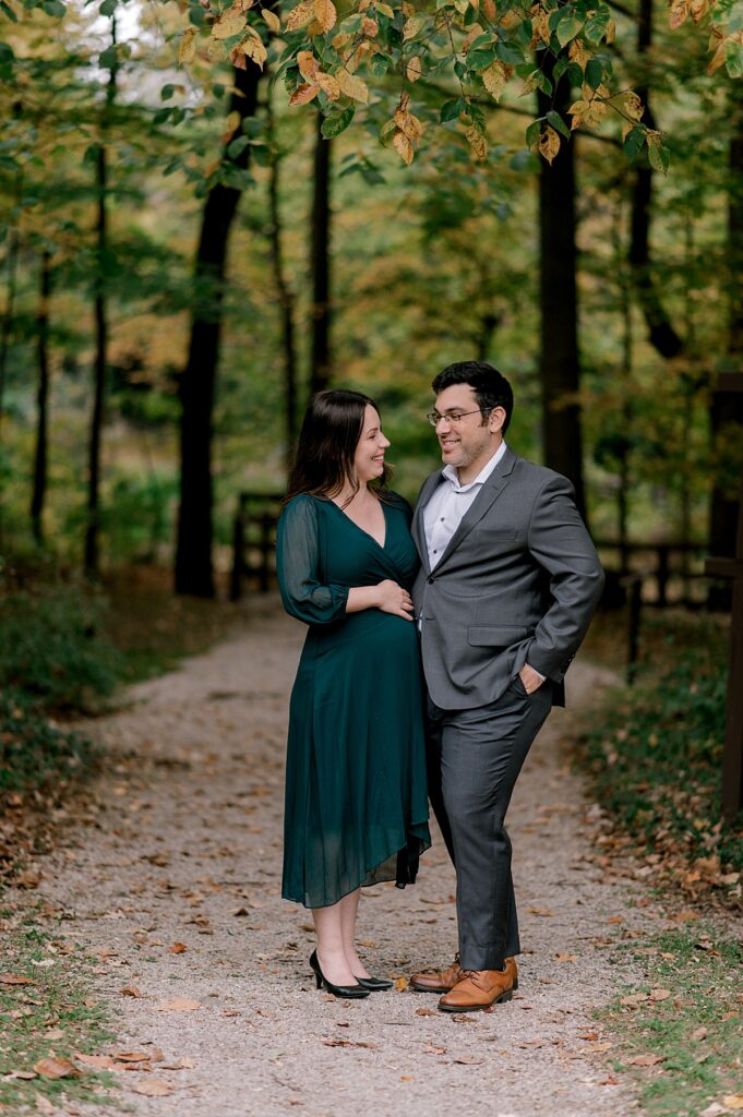 Newly expecting parents standing hip to hip. Mom is in a 3/4 length emerald dress and dad is in a suit jacket. They are smiling and looking at one another while standing under a opening to a gravel path through the woods by North Olmsted Maternity Photographer, Brittany Serowski Photography.