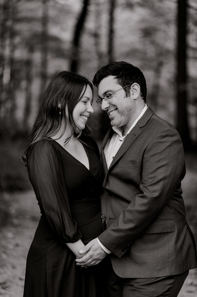A black and white photo of newly expecting parents standing forehead to forehead while holding onto mom's pregnant bump. They are smiling and looking at one another while standing under a opening to a gravel path through the woods by North Olmsted Maternity Photographer, Brittany Serowski Photography.
