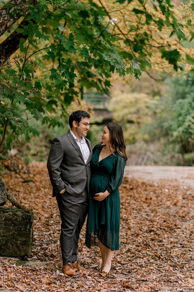 Newly expecting parents standing hip to hip. Mom is in a 3/4 length emerald dress and dad is in a suit jacket. They are smiling and looking at one another while standing under a fall colored tree near a small creek at David Fortier Park by North Olmsted Maternity Photographer, Brittany Serowski Photography.