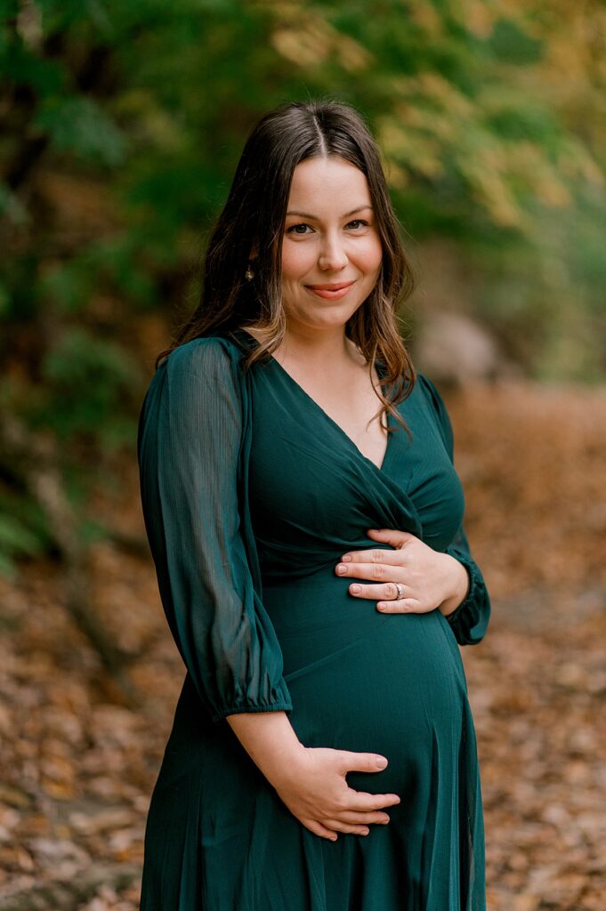 A newly expecting mother wearing a emerald green dress, holding onto her pregnant bump while softly smiling at the camera. She has a fall background behind her at David Fortier Park by North Olmsted Maternity Photographer, Brittany Serowski Photography.