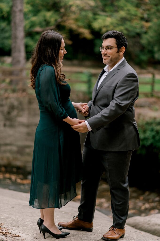Newly expecting parents standing facing one another while holding hands. Mom is in a 3/4 length emerald dress and dad is in a suit jacket. They are smiling and looking at one another while standing next to a small creek at David Fortier Park by North Olmsted Maternity Photographer, Brittany Serowski Photography.