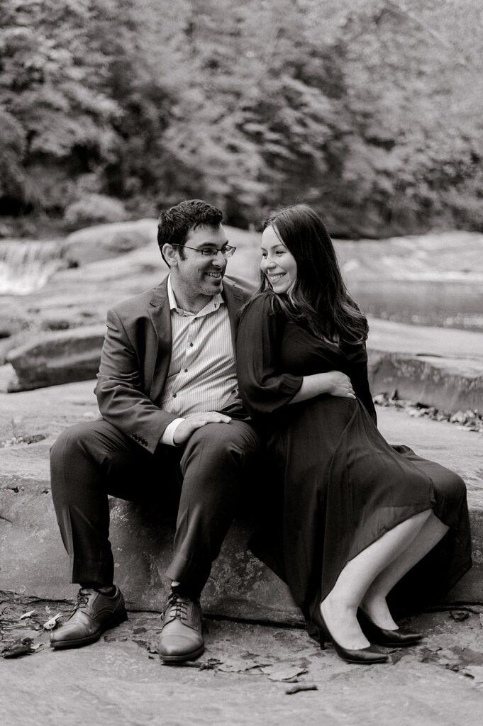 A black and white photo of newly expecting parents sitting side by side on a rock in front of the waterfall at David Fortier Park. Mom is in a dress holding onto her bump and looking over her shoulder at her husband. Dad is in a suit holding onto mom looking and smiling at her by North Olmsted Maternity Photographer, Brittany Serowski Photography.