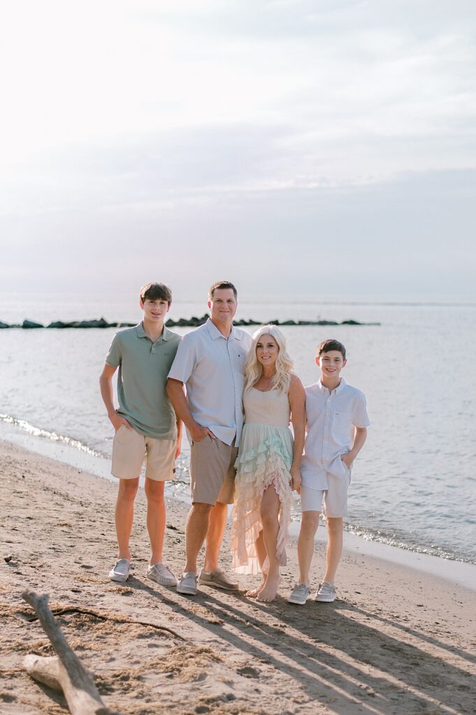 A family of four in light blues, greens and khaki standing together huddled closely in front of Lake Erie for a Vermilion Family Photography session by Brittany Serowski Photography.
