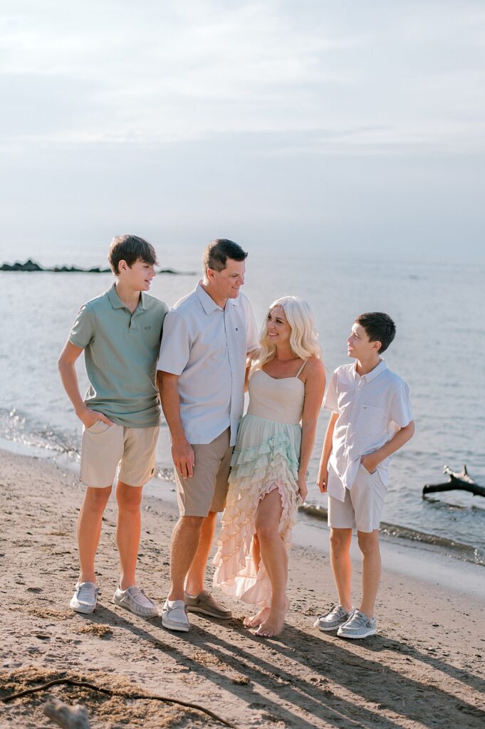 A family of four in light blues, greens and khaki standing together huddled closely in front of Lake Erie, laking at one another and smiling at each other for a Vermilion Family Photography session by Brittany Serowski Photography.