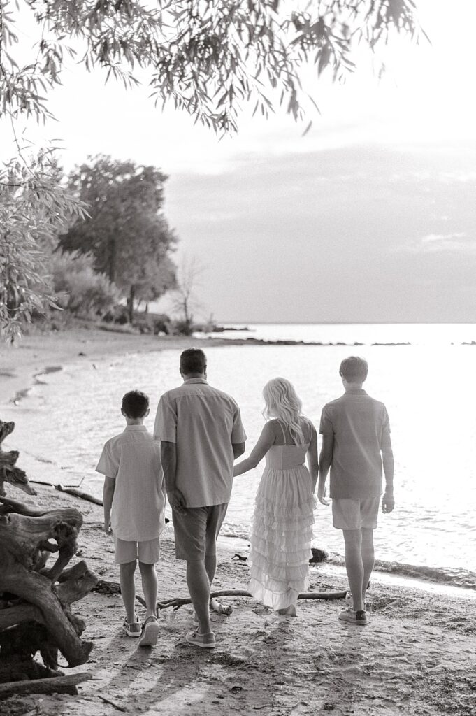A family of four in a black and white photo holding hands, walking away from the camera, along Lake Erie for a Vermilion Family Photography session by Brittany Serowski Photography.