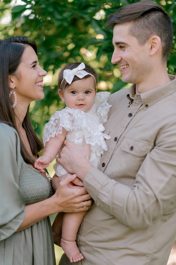 A family of three, with mom and dad holding their daughter in between them while looking at one another and the young baby girl, smiling and looking straight ahead at the camera for a schoepfle garden photography session by Brittany Serowski Photography.