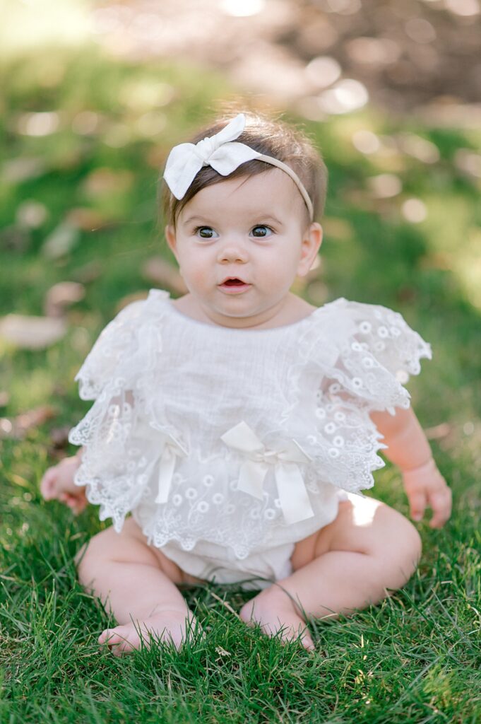 A  young baby girl, in a white lace romper and white headband, sitting on the grass, smiling and looking straight ahead at the camera for a schoepfle garden photography session by Brittany Serowski Photography.