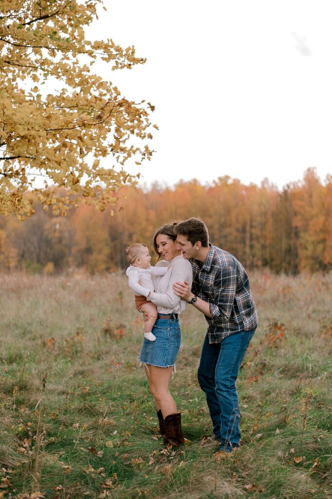 A family of three standing in an open field with their infant baby girl. Mom and dad are smiling at their little girl while mom is holding her. Mom is in a neutral long sleeve shirt and jean skirt. Dad is wearing a flannel button up shirt and dark jeans. A Cleveland Fall Family Photography Session by Brittany Serowski Photography.