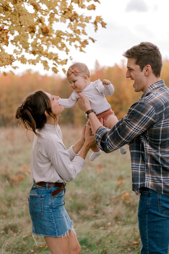A family of three standing in an open field with their infant baby girl. Dad is holding baby girl in the air doing airplane to mom. Mom and baby girl are smiling at each other. Mom is in a neutral long sleeve shirt and jean skirt. Dad is wearing a flannel button up shirt and dark jeans. A Cleveland Fall Family Photography Session by Brittany Serowski Photography.