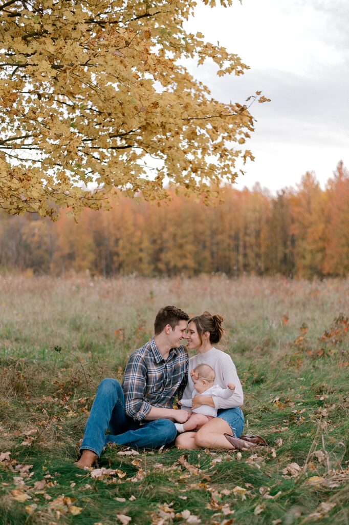 A family of three sitting in an open field with their infant baby girl. Mom and dad are smiling at each other while mom is holding her. Mom is in a neutral long sleeve shirt and jean skirt. Dad is wearing a flannel button up shirt and dark jeans. A Cleveland Fall Family Photography Session by Brittany Serowski Photography.