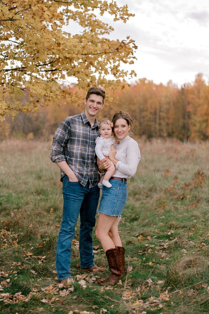 A family of three standing in an open field with their infant baby girl. everyone is smiling at looking at the camera while mom is holding her. Mom is in a neutral long sleeve shirt and jean skirt. Dad is wearing a flannel button up shirt and dark jeans. A Cleveland Fall Family Photography Session by Brittany Serowski Photography.