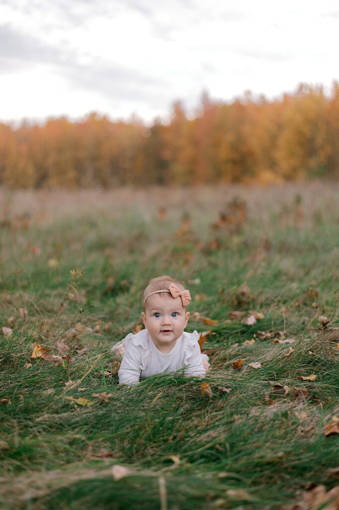 A little baby girl with a white long sleeve shirt on laying in tall grass in a autumn field. She is wearing a peach colored bow headband and smiling towards the camera. A Cleveland Fall Family Photography Session by Brittany Serowski Photography.