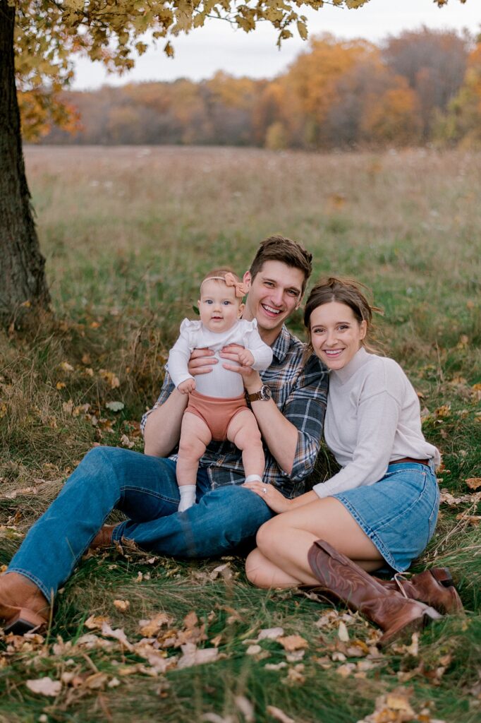 A family of three sitting in an open field with their infant baby girl. everyone is laughing towards the camera while dad is holding baby girl. Mom is in a neutral long sleeve shirt and jean skirt. Dad is wearing a flannel button up shirt and dark jeans. A Cleveland Fall Family Photography Session by Brittany Serowski Photography.