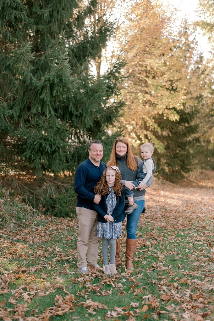 A family of four standing in front of a mix of pine trees and fall colored leaves in gray and navy blue clothing. Everyone is standing close together and smiling at the camera by Bay Village Fall Photographer, Brittany Serowski Photography.