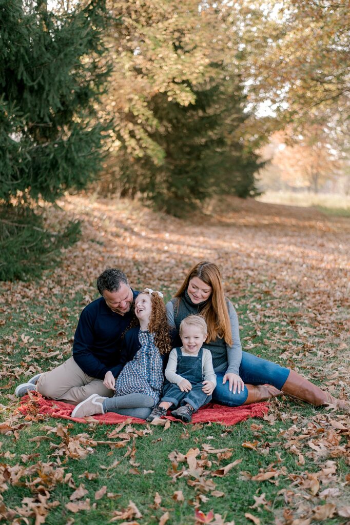 A family of four sitting in front of a mix of pine trees and fall colored leaves in gray and navy blue clothing. Everyone is sitting close together on a blanket and smiling and laughing at one another by Bay Village Fall Photographer, Brittany Serowski Photography.