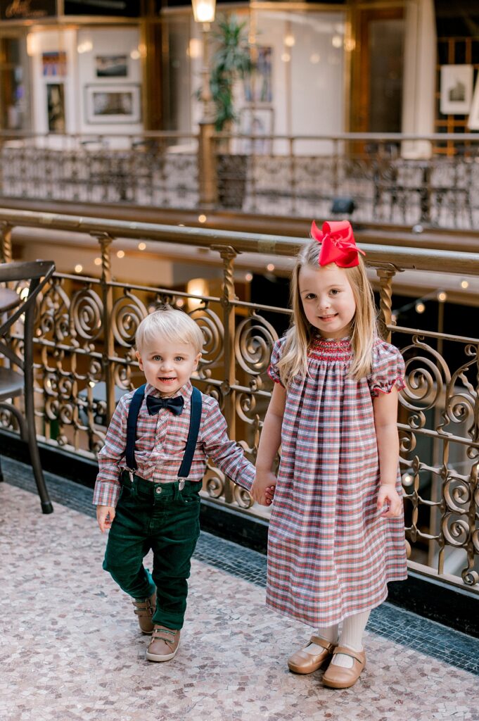 A Cleveland Arcade Family Session by Brittany Serowski Photography. A young sister and brother holding hands in front of a golden iron railing at the Cleveland Arcade. They are wearing matching red and green gingham prints while holding hands and smiling at the camera.