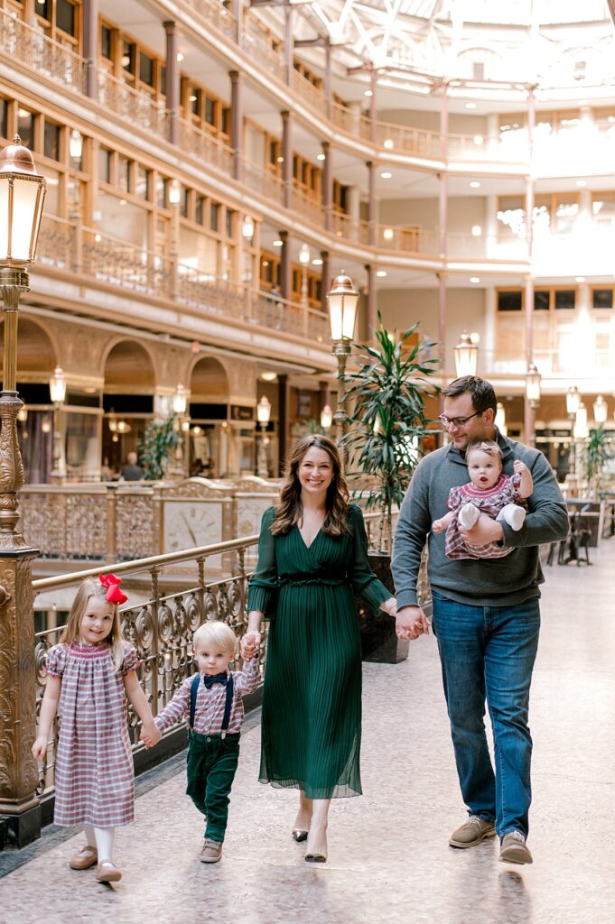 A Cleveland Arcade Family Session by Brittany Serowski Photography. A family of five walking in front of the golden railing and architecture of the Cleveland Arcade in holiday hues of red, green and gray. Dad is holding the young baby girl while looking down at his other two children and holding mom's hand. Mom is smiling at the camera as well as her children as they walk towards the camera.